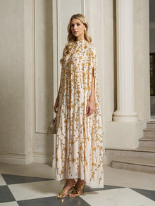 A woman stands indoors wearing the Social Queen Hand-Embellished Floral Caftan Dress, featuring gold embroidery. She pairs it with gold sandals and a small gold handbag, adding elegant resort wear flair against marble floors and columns.
