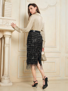 A woman stands indoors facing sideways, wearing a beige blouse, black heels, holding a small handbag, and styled in the Social Queen Hand-Embellished Sequin Fringe Skirt.
