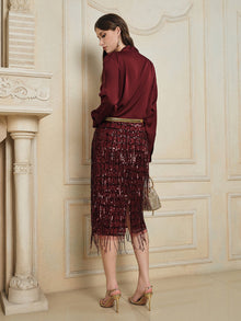 A woman faces away near a decorative wall and fireplace, wearing Social Queen's Hand-Embellished Sequin Fringe Skirt, a burgundy satin blouse, gold heels, and holding a small handbag.