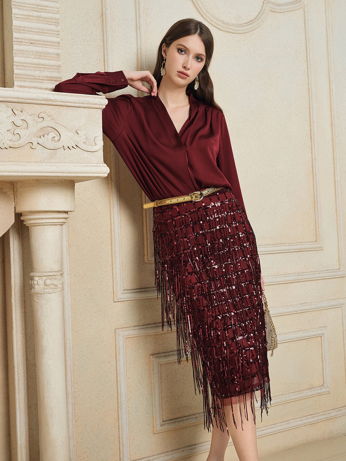A woman in a burgundy blouse and Social Queen’s Hand-Embellished Sequin Fringe Skirt leans against a cream ornate mantel in an elegant room.