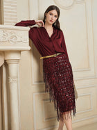 A woman in a burgundy blouse and Social Queen’s Hand-Embellished Sequin Fringe Skirt leans against a cream ornate mantel in an elegant room.