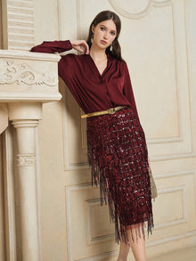 A woman in a burgundy blouse and Social Queen’s Hand-Embellished Sequin Fringe Skirt leans against a cream ornate mantel in an elegant room.