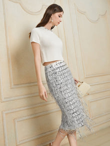 A woman stands sideways in a short-sleeve white top and the Social Queen Hand-Embellished Sequin Fringe Skirt, holding a small beige handbag against an ornate neutral wall background.