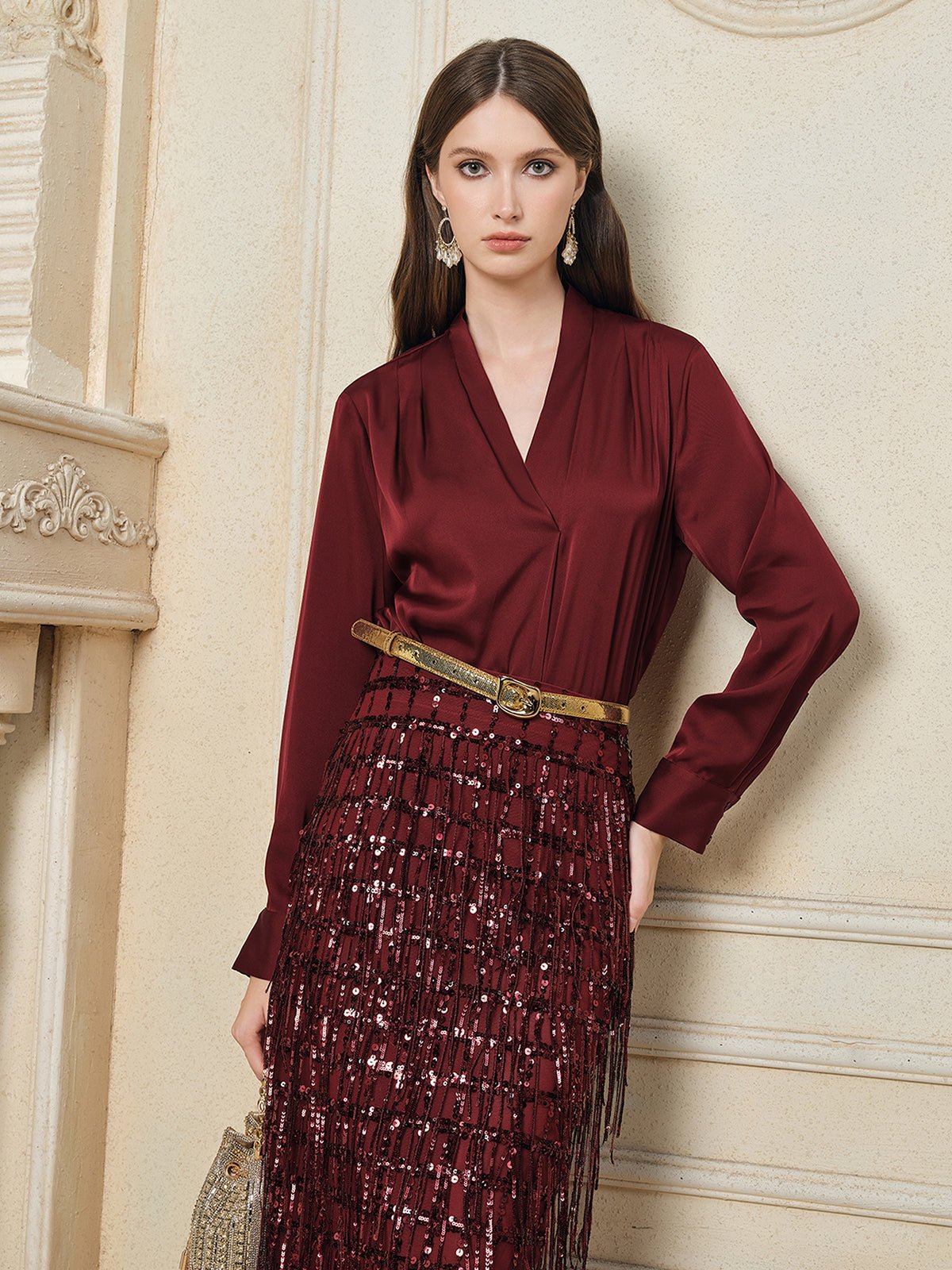 A woman with long brown hair poses against a cream wall, wearing Social Queen’s maroon Hand-Embellished Sequin Fringe Skirt and a matching blouse, accessorized with a small clutch.
