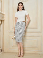 A woman stands indoors against neutral wall paneling, wearing a white short-sleeve textured top and the Social Queen Hand-Embellished Sequin Fringe Skirt in silver, holding a small handbag.
