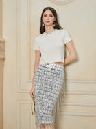 A woman wears the Social Queen Hand-Embellished Sequin Fringe Skirt—a knee-length, high-waisted piece with silver sequin fringe—paired with a short-sleeved white jeweled-neckline top, posed against a cream-colored wall.