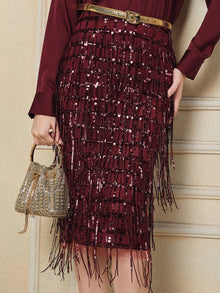 A person wears the Social Queen Hand-Embellished Sequin Fringe Skirt with a gold belt and holds a small, gold beaded handbag against a cream-colored wall.