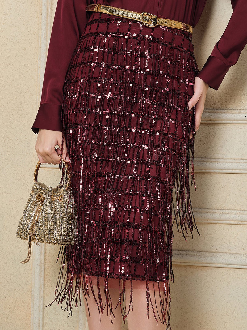 A person wears the Social Queen Hand-Embellished Sequin Fringe Skirt with a gold belt and holds a small, gold beaded handbag against a cream-colored wall.