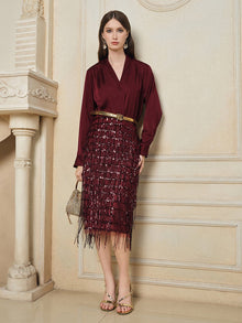 A woman stands indoors wearing a burgundy long-sleeve blouse, gold sandals, statement earrings, and the Social Queen Hand-Embellished Sequin Fringe Skirt with a high waist and fringe details, while holding a small woven handbag.