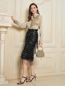 Indoors, a woman wears a beige embroidered blouse, the Social Queen Hand-Embellished Sequin Fringe Skirt, black strappy heels, and carries a small woven handbag.
