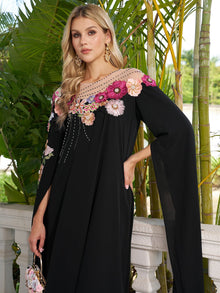 A woman stands outdoors in front of tropical plants, wearing the Social Queen Hand-Embroidered Floral Cape Gown with floral appliqués on the neckline and sleeves, accessorized with gold earrings.