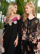 Two women pose outdoors amid lush tropical greenery, both wearing Social Queen’s Hand-Embroidered Floral Cape Gown—showcasing its elegant black hue and exquisite floral details.
