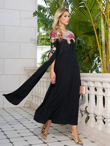 A woman in a Social Queen Hand-Embroidered Floral Cape Gown and gold heels walks on a tiled balcony beside lush green plants.