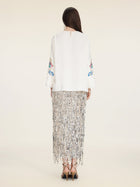 A woman stands facing away, wearing the Social Queen Hand-Embroidered Floral Jacquard Pullover with floral sleeve motifs, a silver fringe skirt, and black heeled sandals.