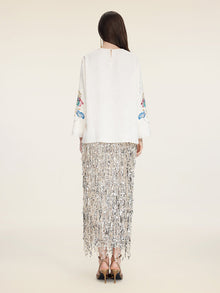 A woman stands facing away, wearing the Social Queen Hand-Embroidered Floral Jacquard Pullover with floral sleeve motifs, a silver fringe skirt, and black heeled sandals.