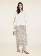 A woman stands against a neutral background wearing the Social Queen Hand-Embroidered Floral Jacquard Pullover, paired with a silver fringe skirt, gold high-heeled sandals, and a small metallic handbag.