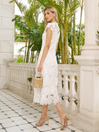 A woman in the Social Queen Hand-Embroidered Floral Lace Dress, gold heels, and a woven bag stands on a balcony with tropical plants—the perfect stylish wedding guest look.