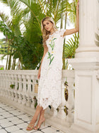 A woman in the Social Queen Hand-Embroidered Floral Lace Dress with gold heels stands by a white balustrade and lush tropical plants—a graceful choice for a wedding guest.