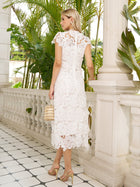A woman in the Social Queen Hand-Embroidered Floral Lace Dress with heels and a small handbag stands on a tiled balcony amid greenery—the ideal look for a wedding guest.