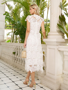 A woman in the Social Queen Hand-Embroidered Floral Lace Dress with heels and a small handbag stands on a tiled balcony amid greenery—the ideal look for a wedding guest.