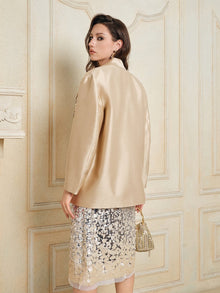 A woman faces backward in a decorated beige room, wearing the Social Queen Hand-Embroidered Floral Silk-Wool Jacket over a sequin skirt and holding a small beaded handbag.