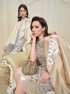 Two women in Hand-Embroidered Floral Silk-Wool Jackets by Social Queen sit on an ornate cream chair, styled with sequined skirts and gold accessories, both in chic champagne satin blazers with pearl buttons.