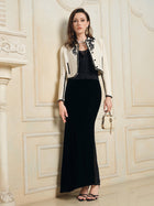 A woman stands indoors against a decorated wall, wearing Social Queen’s Hand-Embroidered Silk-Wool Cropped Tweed Jacket with black embroidery, paired with a black top, long black skirt, sandals, and a small gold handbag.