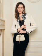 A woman stands indoors wearing Social Queen's Hand-Embroidered Silk-Wool Cropped Tweed Jacket with black trim over a black dress, holding a cream handbag and accessorized with jewelry.