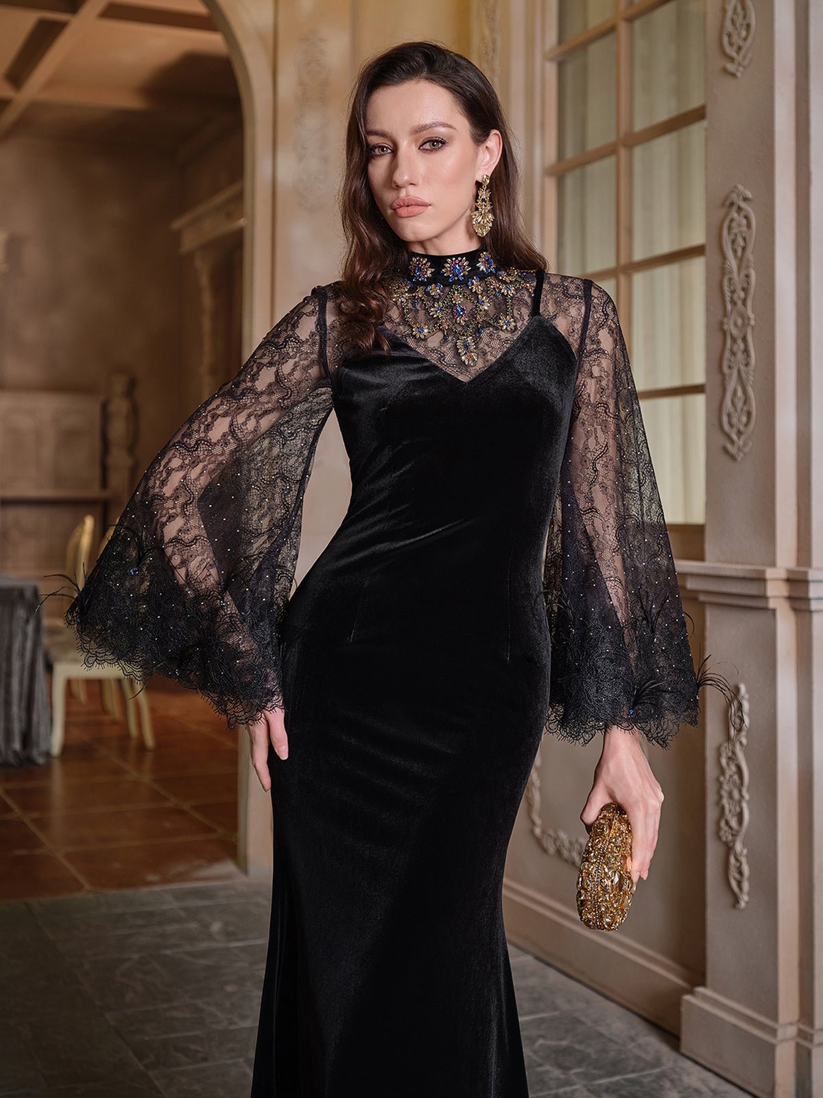 A woman in the Social Queen Hand-Sewn Crystal Velvet Feather Mermaid Gown stands indoors, holding a gold clutch and looking at the camera.