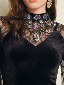 A woman wears the Social Queen Hand-Sewn Crystal Velvet Feather Mermaid Gown, a black velvet dress with lace details, mermaid silhouette, high collar, and colorful crystal embellishments.