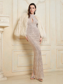 A woman models Social Queen's Hand-Sewn Pearl & Sequin Halter Gown and a white fur jacket, posing indoors against an ornate, cream-colored wall.