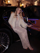 A woman in the back seat of a luxury car at night wears the Social Queen Hand-Sewn Pearl & Sequin Halter Gown and a white fur coat, surrounded by festive lights and decorations.