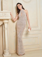 A woman stands indoors, leaning against a mantelpiece and holding a clutch, wearing the Social Queen Hand-Sewn Pearl & Sequin Halter Gown with a sleeveless, floor-length silhouette and a shimmering sequin skirt.