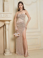 A woman indoors wears the Social Queen Hand-Sewn Radiant Crystal Mesh Pleated Gown, featuring a strapless design and high slit, paired with a floral handbag and high-heeled sandals.