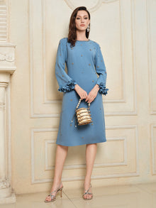 A woman wears the Social Queen Handcrafted Beaded Floral A-Line Dress with ruffled sleeves, holding a small gold handbag, standing against a cream wall.