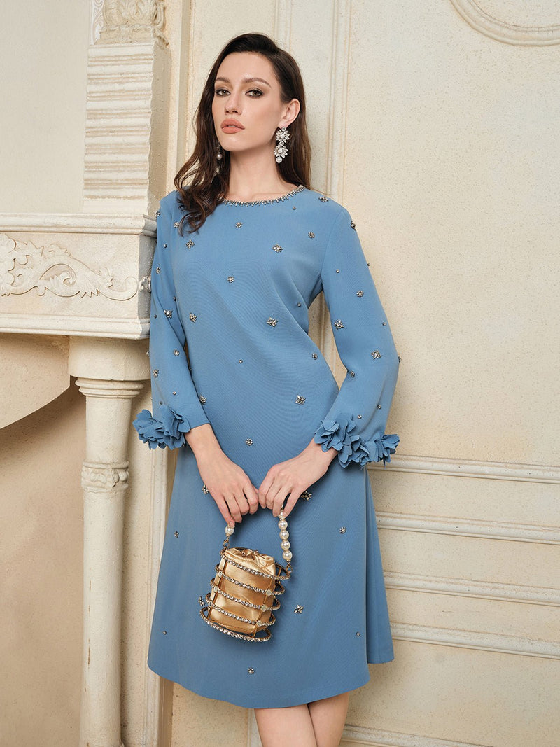 A woman wears the Social Queen Handcrafted Beaded Floral A-Line Dress, featuring blue long sleeves and floral embellishments, while holding a small structured handbag indoors against a cream-colored wall.
