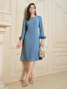 A woman in a Social Queen Handcrafted Beaded Floral A-Line Dress stands indoors against a beige wall, carrying a small handbag and wearing silver strappy heels.