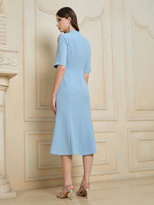 A woman in the Social Queen Handcrafted Beaded Floral Dress, sky-blue and knee-length with short sleeves and 3D floral appliqués, stands indoors facing away in strappy high heels.