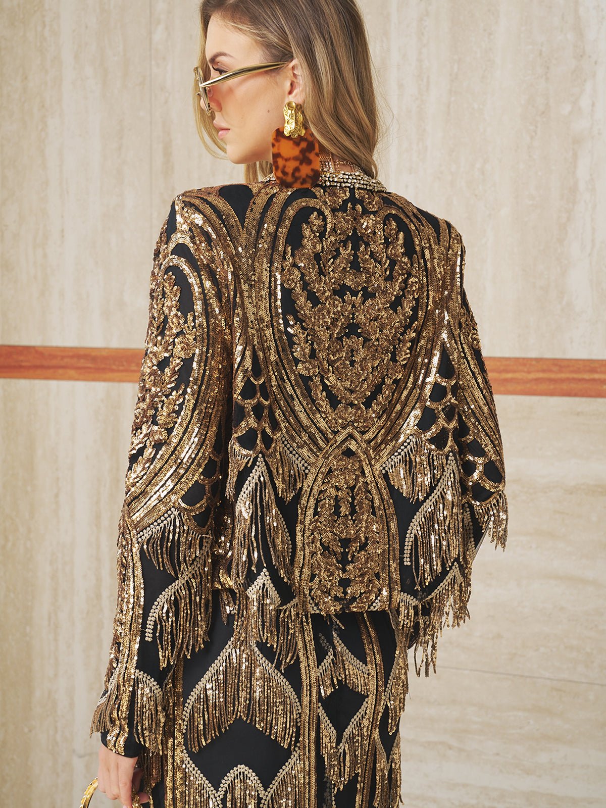 A woman in large tortoiseshell earrings and sunglasses wears the Social Queen Handcrafted Beaded Fringe Jacket, featuring black and gold beading with intricate embroidery and fringe, standing against a beige wall.