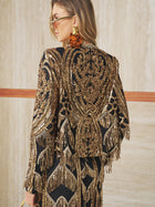 A woman in large tortoiseshell earrings and sunglasses wears the Social Queen Handcrafted Beaded Fringe Jacket, featuring black and gold beading with intricate embroidery and fringe, standing against a beige wall.