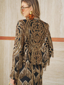 A woman in large tortoiseshell earrings and sunglasses wears the Social Queen Handcrafted Beaded Fringe Jacket, featuring black and gold beading with intricate embroidery and fringe, standing against a beige wall.