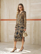 Woman in a black dress with gold details, gold earrings, sunglasses, and heels stands indoors holding a small gold handbag topped with the Social Queen Handcrafted Beaded Fringe Jacket.
