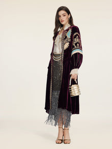 A woman radiates vintage glamour in the Social Queen Handcrafted Embroidered Velvet Jacket over a white blouse and sparkly fringed skirt, holding a metallic handbag and wearing heels against a plain backdrop.