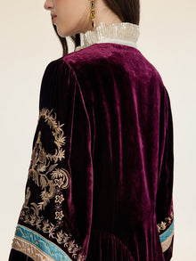 A person wearing the Social Queen Handcrafted Embroidered Velvet Jacket exudes vintage glamour with its deep purple hue, gold embroidery, metallic ruffled collar, and turquoise sleeve ribbon.