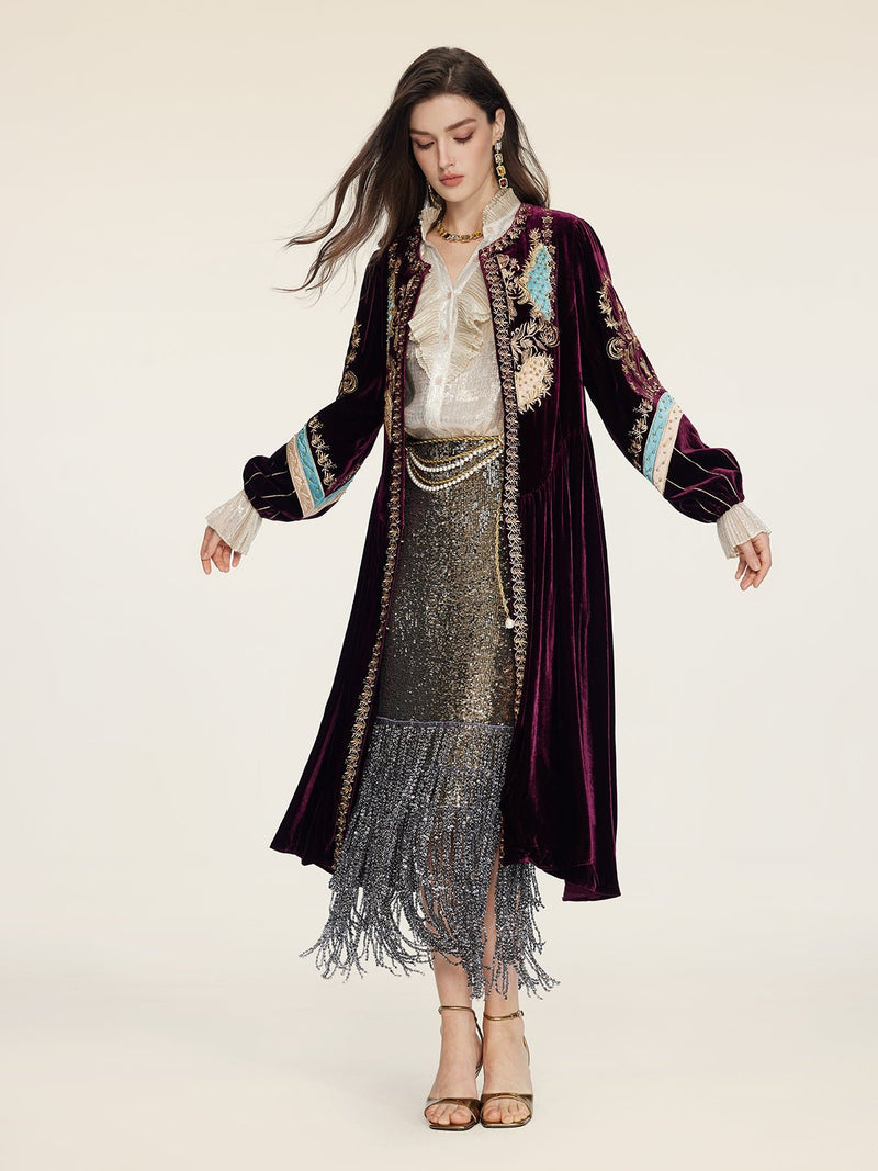 A woman stands against a plain background wearing the Social Queen Handcrafted Embroidered Velvet Jacket over a metallic blouse and fringed sequined skirt with high-heeled sandals, radiating vintage glamour.