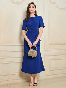 A woman wears the Social Queen Handcrafted Gold-Trimmed Slit Dress, paired with a gold beaded handbag, standing against a cream-colored wall with decorative molding.