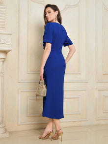 A woman stands before a cream paneled wall in Social Queen’s Handcrafted Gold-Trimmed Slit Dress, paired with gold heels and earrings, holding a small handbag.