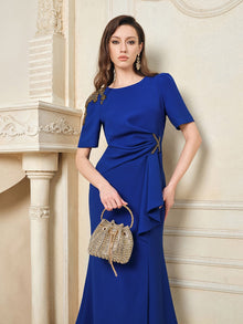 A woman stands indoors in the Social Queen Handcrafted Gold-Trimmed Slit Dress, holding a beige handbag with a round handle.