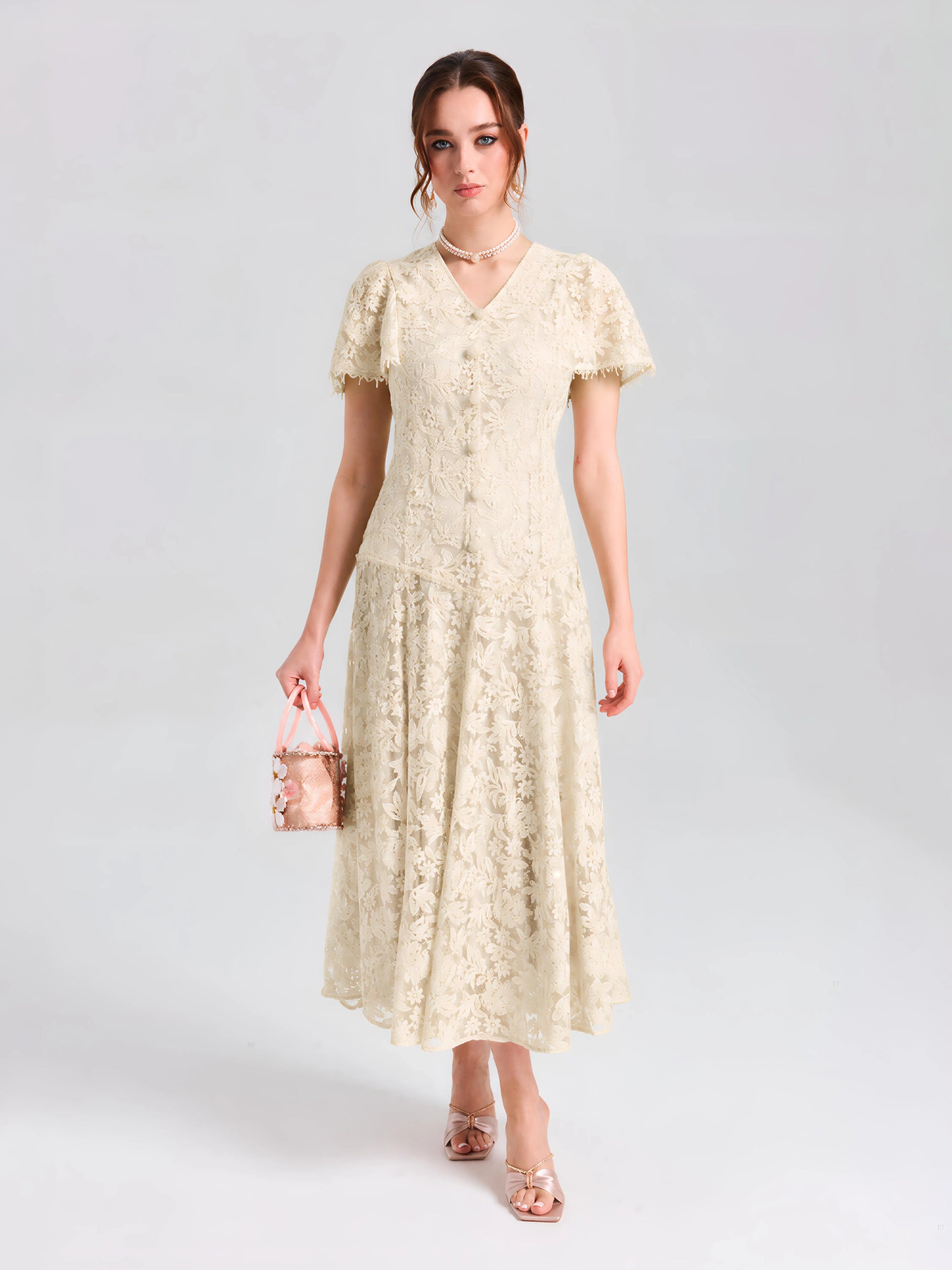 A woman wears the Social Queen Floral Embroidered Lace Dress—a cream A-line midi with short sleeves—paired with a small pink handbag, set against a plain light background.