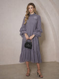 A woman holds a black purse while wearing the Social Queen Floral Appliqué Embroidered Ruffle Dress, featuring long sleeves and delicate details, standing against a beige textured wall.